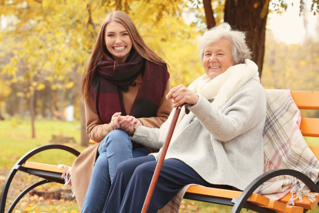 Senior woman with cane and young caregiver sitting on bench in park