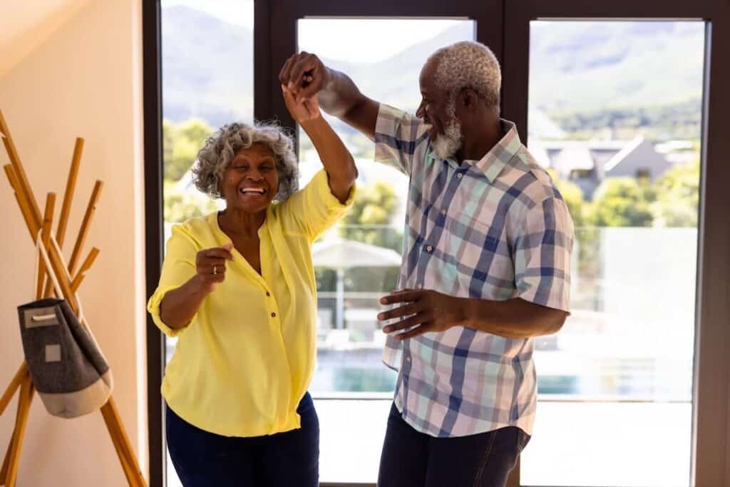 Cheerful african american senior man doing salsa dance with woman against window